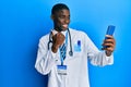 Young african american man wearing doctor uniform using smartphone pointing thumb up to the side smiling happy with open mouth Royalty Free Stock Photo