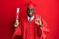 Young african american man wearing graduation cap and ceremony robe holding diploma doing ok sign with fingers, smiling friendly Royalty Free Stock Photo