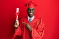 Young african american man wearing graduation cap and ceremony robe holding diploma smiling happy pointing with hand and finger Royalty Free Stock Photo