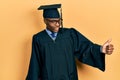 Young african american man wearing graduation cap and ceremony robe looking proud, smiling doing thumbs up gesture to the side Royalty Free Stock Photo