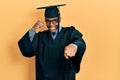 Young african american man wearing graduation cap and ceremony robe smiling doing talking on the telephone gesture and pointing to Royalty Free Stock Photo