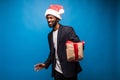 Young African American man wearing a santa hat offering a gift , isolated on blue background Royalty Free Stock Photo