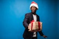 Young African American man wearing a santa hat offering a gift , isolated on blue background Royalty Free Stock Photo