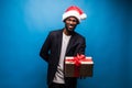 Young African American man wearing a santa hat offering a gift , isolated on blue background Royalty Free Stock Photo