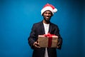 Young African American man wearing a santa hat offering a gift , isolated on blue background Royalty Free Stock Photo