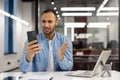 A young African American man works in the office sitting worried at the desk and looking at the phone screen in Royalty Free Stock Photo