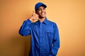 Young african american mechanic man wearing blue uniform and cap over yellow background smiling doing phone gesture with hand and Royalty Free Stock Photo