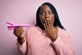 Young african american plus size woman with braids holding pink paper airplane cover mouth with hand shocked with shame for