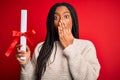 Young african american student girl holding university diploma over red isolated background cover mouth with hand shocked with Royalty Free Stock Photo