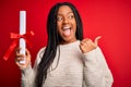 Young african american student girl holding university diploma over red isolated background pointing and showing with thumb up to Royalty Free Stock Photo