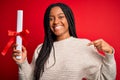 Young african american student girl holding university diploma over red isolated background with surprise face pointing finger to Royalty Free Stock Photo