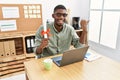 Young african american student man holding graduate degree diploma sitting on the table pointing thumb up to the side smiling