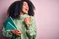 Young african american student woman with afro hair reading books over pink background pointing and showing with thumb up to the Royalty Free Stock Photo