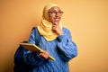 Young African American student woman wearing muslim hijab and backpack holding book with hand on chin thinking about question, Royalty Free Stock Photo