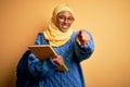 Young African American student woman wearing muslim hijab and backpack holding book pointing to you and the camera with fingers, Royalty Free Stock Photo