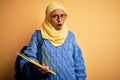 Young African American student woman wearing muslim hijab and backpack holding book In shock face, looking skeptical and