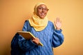 Young African American student woman wearing muslim hijab and backpack holding book Waiving saying hello happy and smiling,