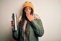 Young african american tourtist woman wearing coat and drinking water from thermo cover mouth with hand shocked with shame for Royalty Free Stock Photo