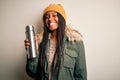 Young african american tourtist woman wearing coat and drinking water from thermo with a happy face standing and smiling with a Royalty Free Stock Photo