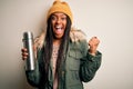 Young african american tourtist woman wearing coat and drinking water from thermo screaming proud and celebrating victory and Royalty Free Stock Photo