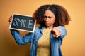 Young african american woman with afro hair holding blackboard with smile message pointing with finger to the camera and to you, Royalty Free Stock Photo