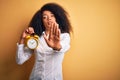 Young african american woman with afro hair holding classic alarm clock over yellow background with open hand doing stop sign with Royalty Free Stock Photo
