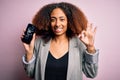 Young african american woman with afro hair holding cracked and broken smartphone screen doing ok sign with fingers, excellent