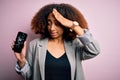 Young african american woman with afro hair holding cracked and broken smartphone screen stressed with hand on head, shocked with