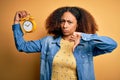 Young african american woman with afro hair holding vintage alarm clock over yellow background with angry face, negative sign Royalty Free Stock Photo