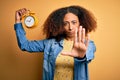 Young african american woman with afro hair holding vintage alarm clock over yellow background with open hand doing stop sign with Royalty Free Stock Photo