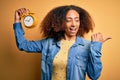 Young african american woman with afro hair holding vintage alarm clock over yellow background pointing and showing with thumb up Royalty Free Stock Photo