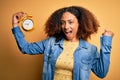 Young african american woman with afro hair holding vintage alarm clock over yellow background screaming proud and celebrating Royalty Free Stock Photo