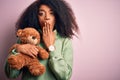 Young african american woman with afro hair hugging teddy bear over pink background cover mouth with hand shocked with shame for Royalty Free Stock Photo