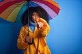 Young african american woman with afro hair under colorful umbrella wearing winter coat for rain pointing with finger to the Royalty Free Stock Photo