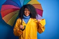 Young african american woman with afro hair under colorful umbrella wearing winter coat for rain with surprise face pointing Royalty Free Stock Photo