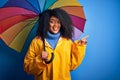Young african american woman with afro hair under colorful umbrella wearing winter coat for rain very happy pointing with hand and Royalty Free Stock Photo