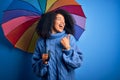 Young african american woman with afro hair under colorful umbrella for winter weather rain pointing and showing with thumb up to Royalty Free Stock Photo