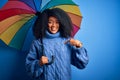 Young african american woman with afro hair under colorful umbrella for winter weather rain with surprise face pointing finger to Royalty Free Stock Photo