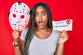 Young african american woman with braids holding halloween banner and scary mask afraid and shocked with surprise and amazed