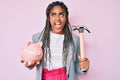 Young african american woman with braids holding piggy bank and hammer angry and mad screaming frustrated and furious, shouting Royalty Free Stock Photo