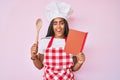 Young african american woman with braids wearing professional baker apron reading cooking recipe book winking looking at the Royalty Free Stock Photo