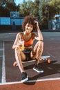 young african-american woman in bright sports bra sitting on longboard at sport ground with orange juice