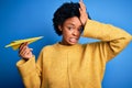 Young African American woman with curly hair holding paper airplane over blue background stressed with hand on head, shocked with Royalty Free Stock Photo