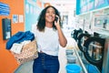 Young african american woman with curly hair smiling happy doing chores at the laundry speaking on the phone Royalty Free Stock Photo