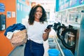 Young african american woman with curly hair smiling happy doing chores at the laundry using phone Royalty Free Stock Photo