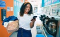 Young african american woman with curly hair smiling happy doing chores at the laundry using phone Royalty Free Stock Photo