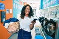 Young african american woman with curly hair smiling happy doing chores at the laundry using phone Royalty Free Stock Photo