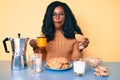 Young african american woman eating breakfast holding croissant making fish face with mouth and squinting eyes, crazy and comical
