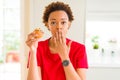 Young african american woman eating chocolate chips muffins cover mouth with hand shocked with shame for mistake, expression of Royalty Free Stock Photo