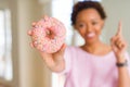 Young african american woman eating pink sugar donut surprised with an idea or question pointing finger with happy face, number Royalty Free Stock Photo